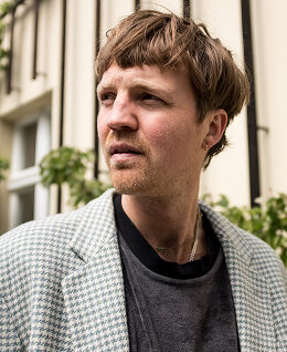 Paul-img - URBAN UTOPIA Mann mit kurzem, lockigem Haar und einem gestreiften Blazer, schaut nachdenklich zur Seite vor einer urbanen Kulisse.