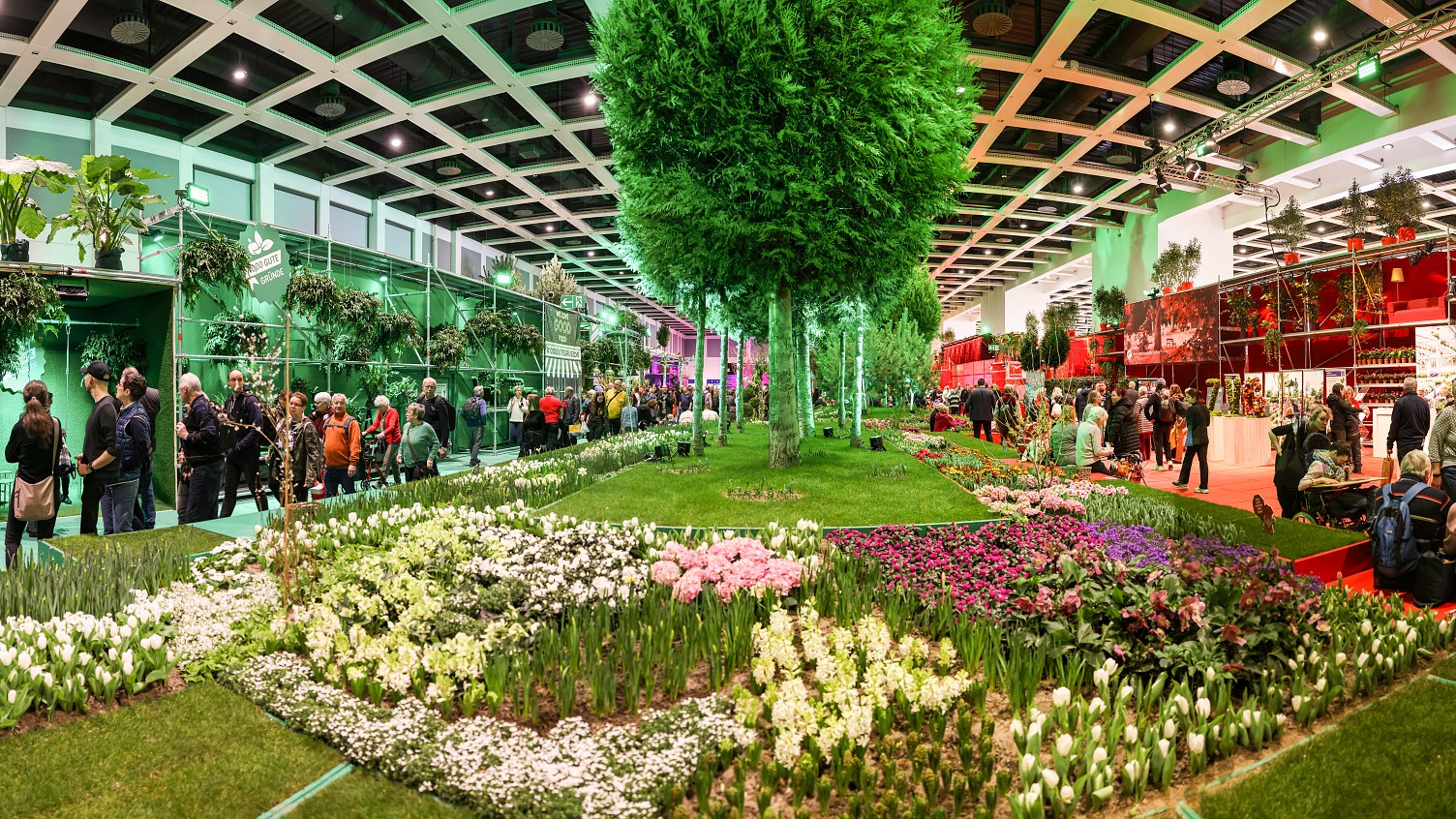 thementour-garten_edited - URBAN UTOPIA Bunte Blumenbeete und große Bäume in einer Messehalle, Besucher schlendern durch die Gartenlandschaft.